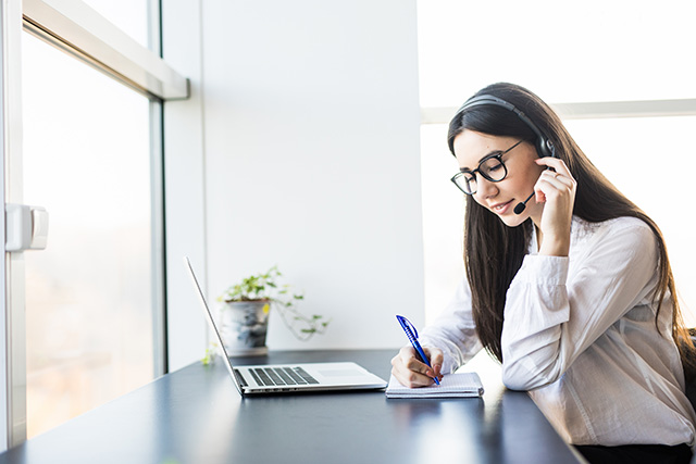 Receptionist aligning headset while taking notes next to her laptop