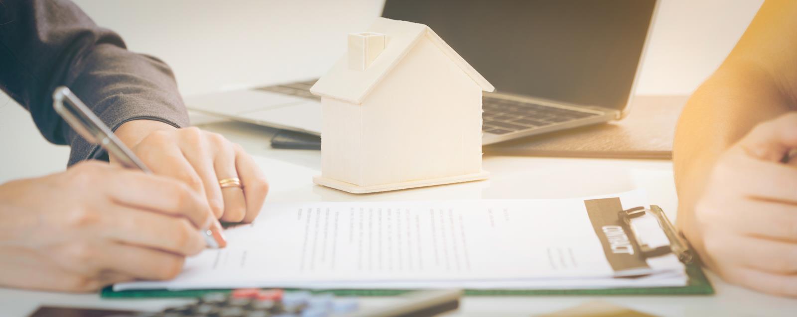 image of a set of two pairs of hands one writing another waiting on a contract or document with a toy house on top of clipboard of paperwork